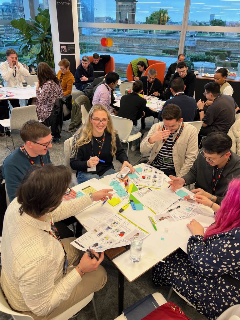A group of people at Primark's ideation workshop gathered around tbales, collaboratively creating paper prototypes. The participants are using materials like paper, scissors, glue and pens, fostering a creative and interactive atmosphere. 